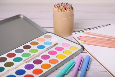 Watercolor palette with brush, pencils, markers and notebook on white wooden table, closeup