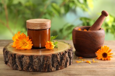 Photo of Jar of cosmetic product, mortar and beautiful calendula flowers on wooden table outdoors