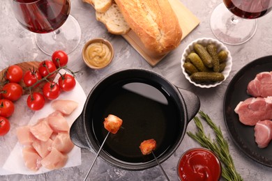 Fondue pot, forks with fried meat pieces, glasses of red wine and other products on grey textured table, flat lay