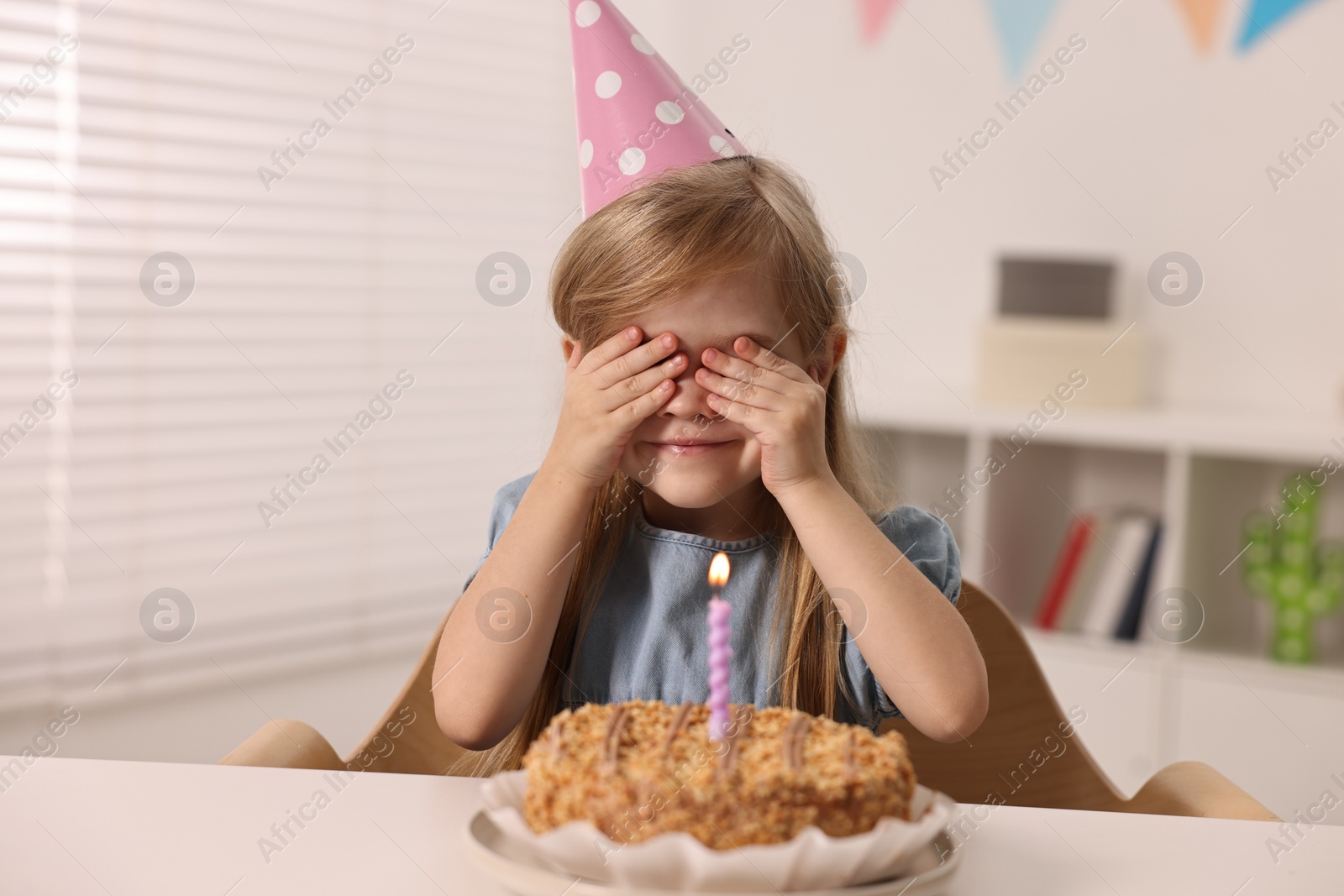 Photo of Cute girl in party hat with birthday cake at table indoors