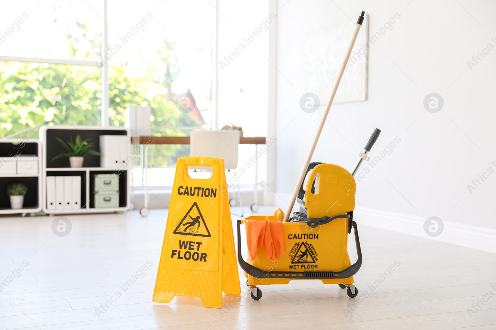 Photo of Phrase "CAUTION WET FLOOR" on safety sign and mop bucket with cleaning supplies, indoors