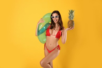 Photo of Beautiful woman in stylish bikini with pineapple and inflatable ring on yellow background