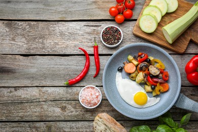 Frying pan with tasty egg and vegetables near ingredients on wooden table, flat lay. Space for text