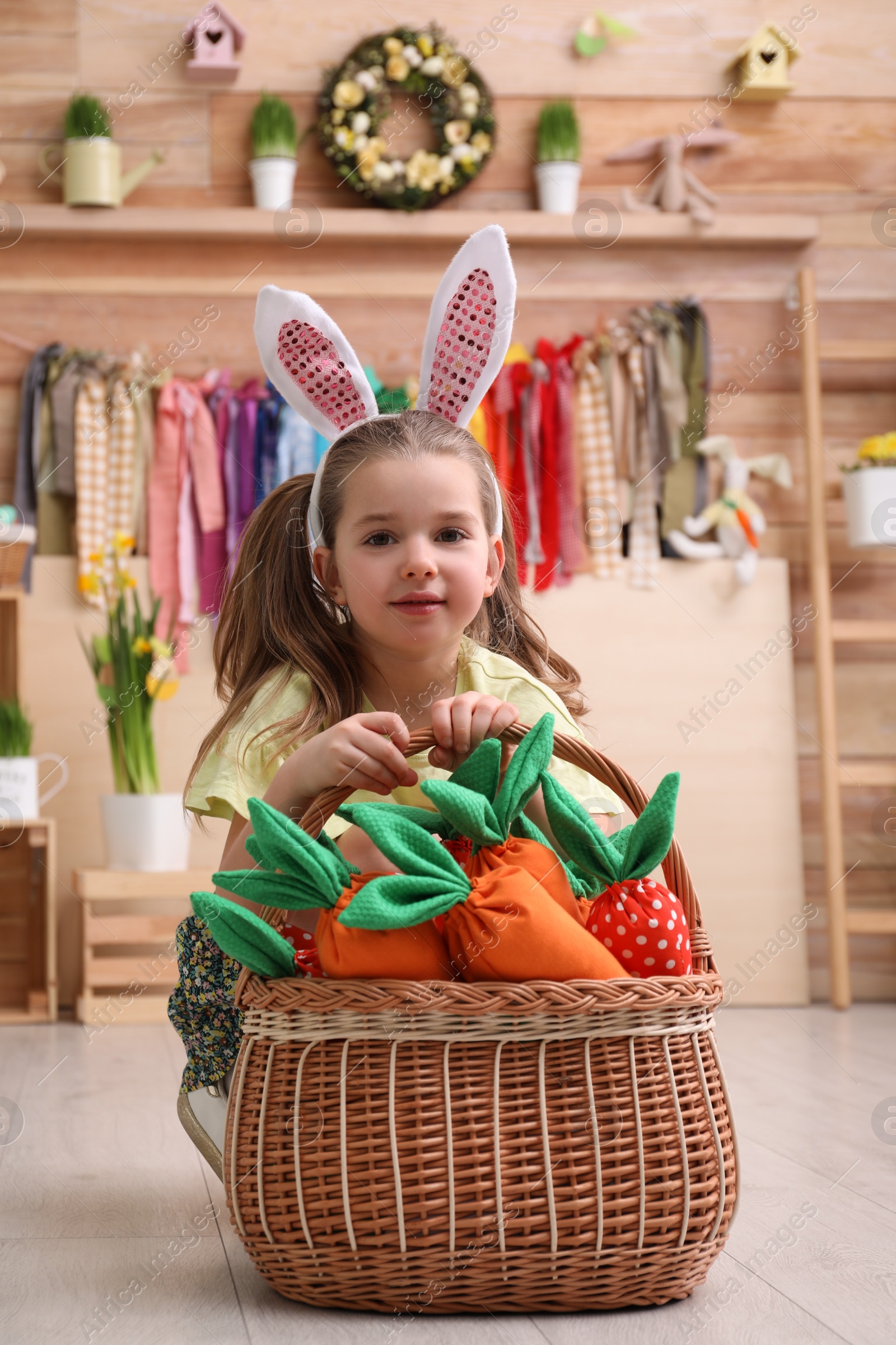 Photo of Adorable little girl with bunny ears and basket full of toy carrots in Easter photo zone