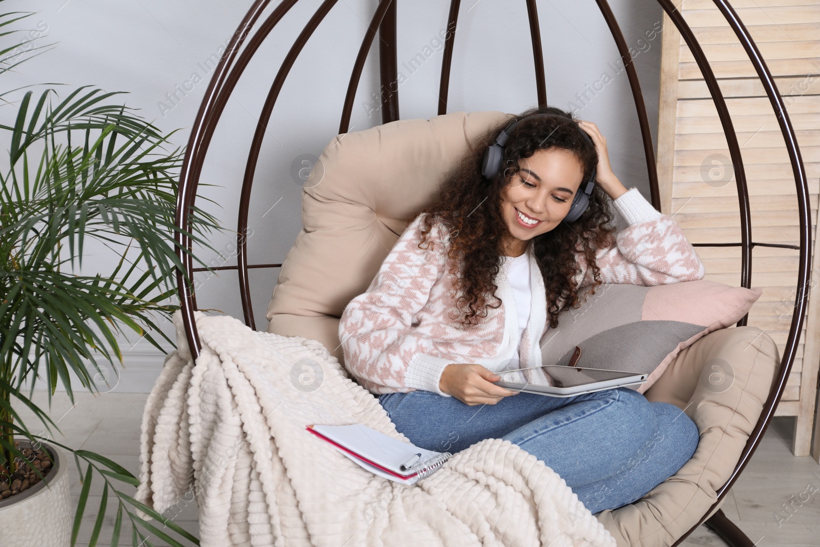 Photo of African American woman with headphones and tablet studying in egg chair at home. Distance learning