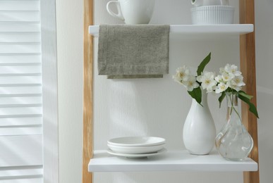Beautiful jasmine flowers and tableware on shelving unit in kitchen