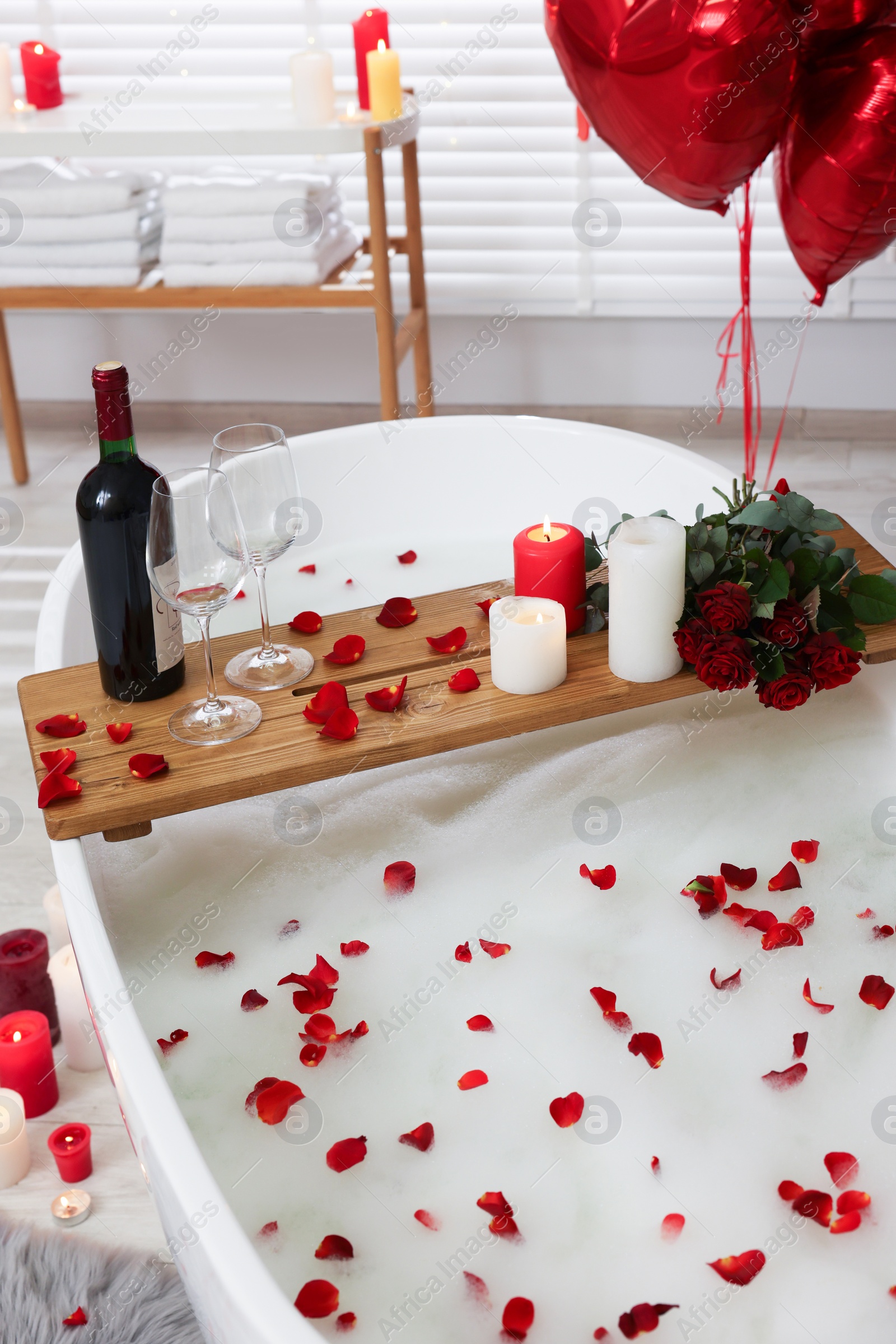 Photo of Wooden tray with wine, burning candles and rose petals on tub in bathroom. Valentine's day celebration