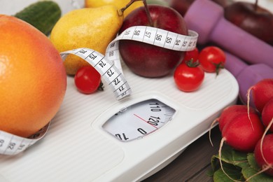 Photo of Scales, healthy food, measuring tape and dumbbells on wooden table, closeup