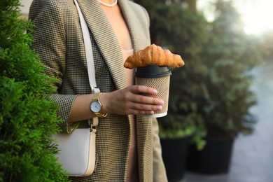 Photo of Woman holding tasty croissant and cup of coffee outdoors, closeup