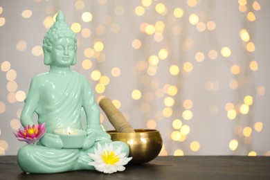 Photo of Buddha statue with candle, lotus flowers and singing bowl on table against blurred lights. Space for text