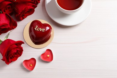 St. Valentine's Day. Delicious heart shaped cake, tea, roses and candles on white wooden table, flat lay. Space for text