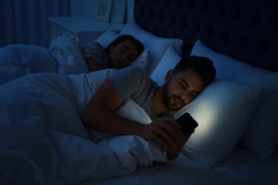 Photo of Young man using smartphone while his girlfriend sleeping in bed at night