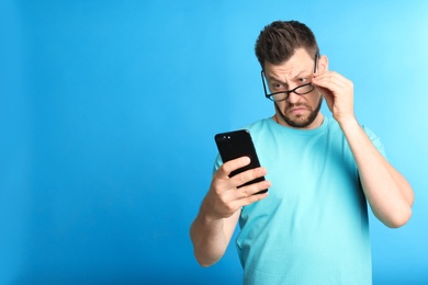 Man with vision problems using smartphone on blue background, space for text