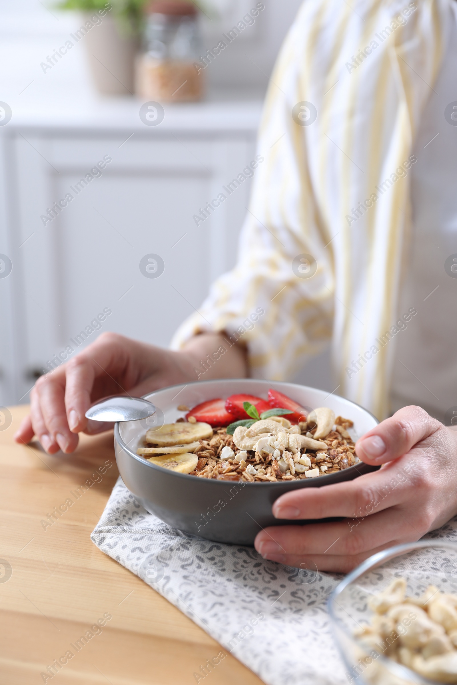 Photo of Woman eating tasty granola with banana, cashew and strawberries at wooden table indoors, closeup