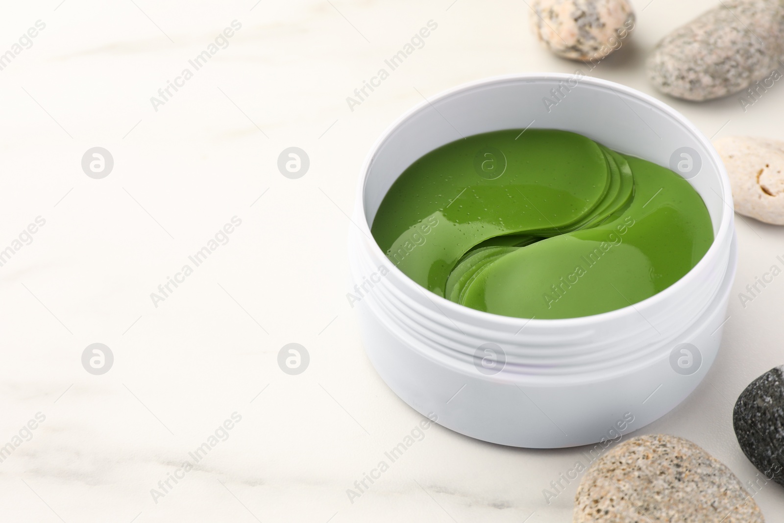 Photo of Jar of under eye patches and stones on white marble table, closeup with space for text. Cosmetic product