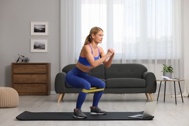 Photo of Athletic woman doing squats with fitness elastic band near laptop on mat at home
