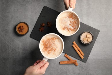 Women holding glass cups of delicious eggnog with cinnamon and anise at grey table, top view