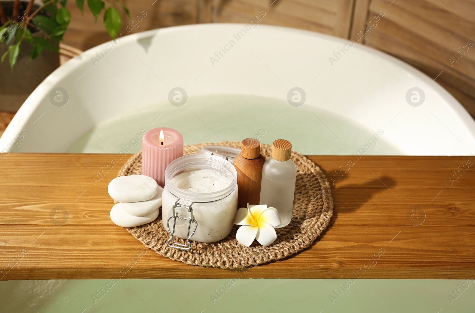 Photo of Wooden tray with spa products, stones and burning candle on bath tub in bathroom