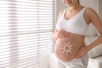 Young pregnant woman with sun protection cream on belly near window, closeup. Space for text
