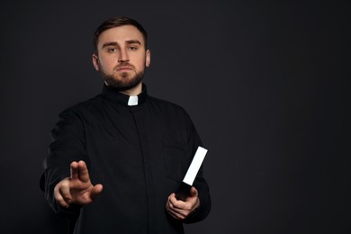 Priest with Bible making blessing gesture on black background. Space for text