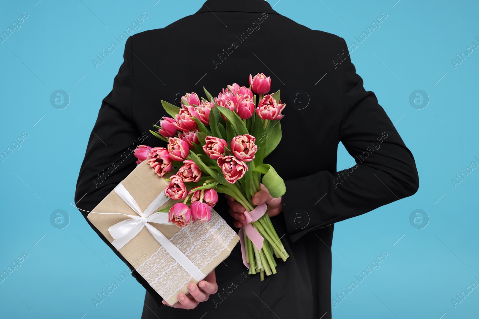 Photo of Man hiding beautiful bouquet and present on light blue background, back view