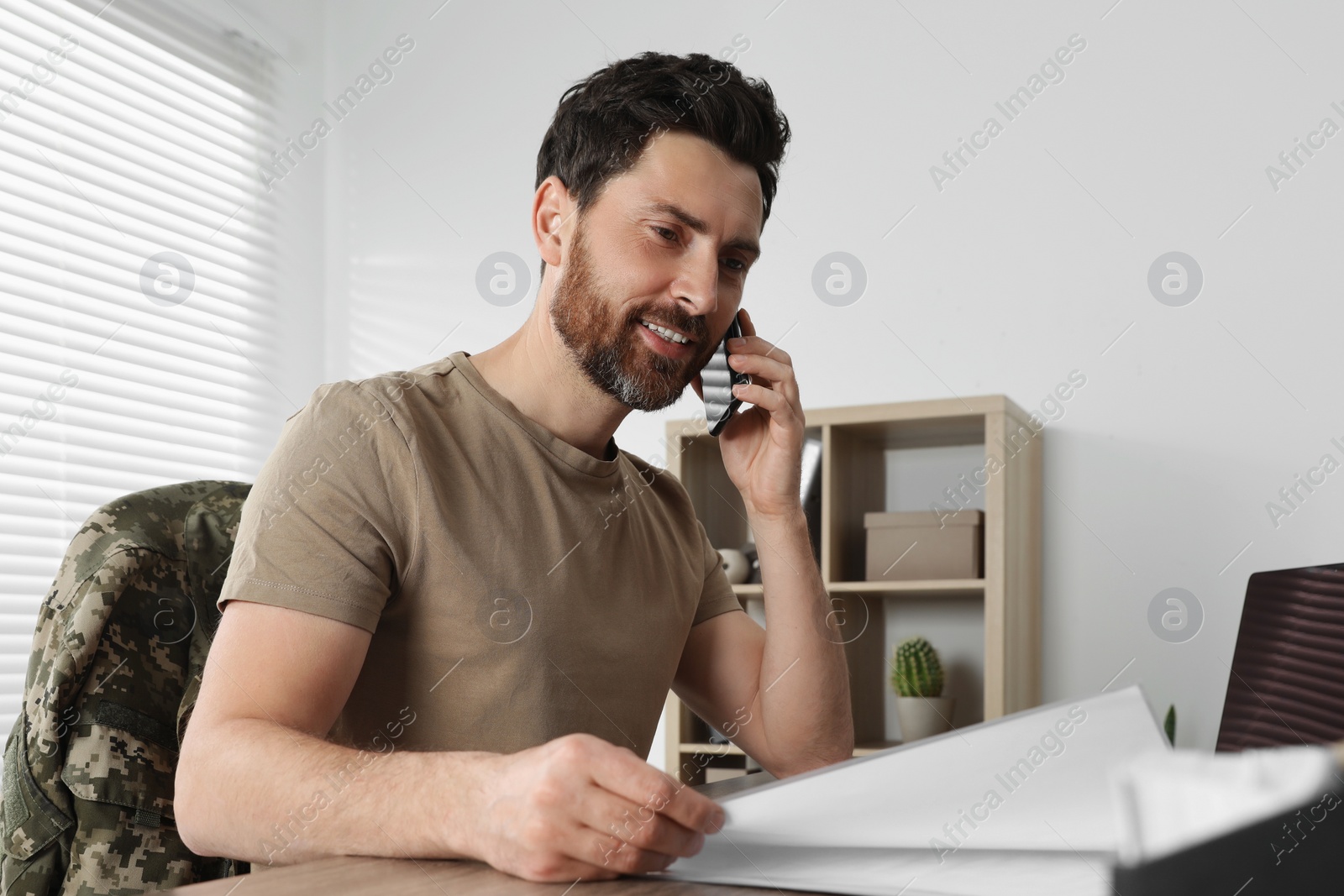 Photo of Happy soldier talking on phone at wooden table indoors. Military service