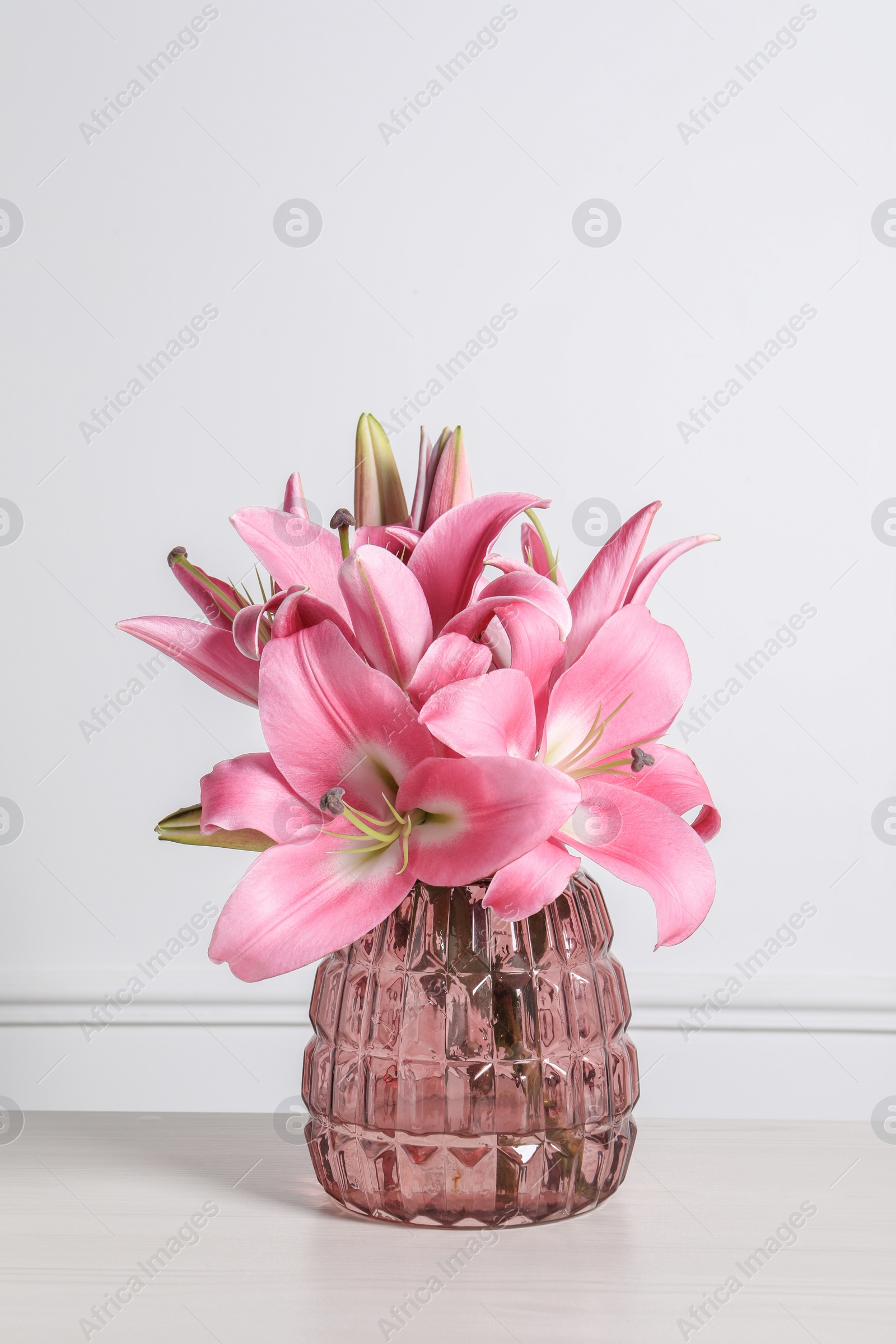 Photo of Beautiful pink lily flowers in vase on wooden table against white wall