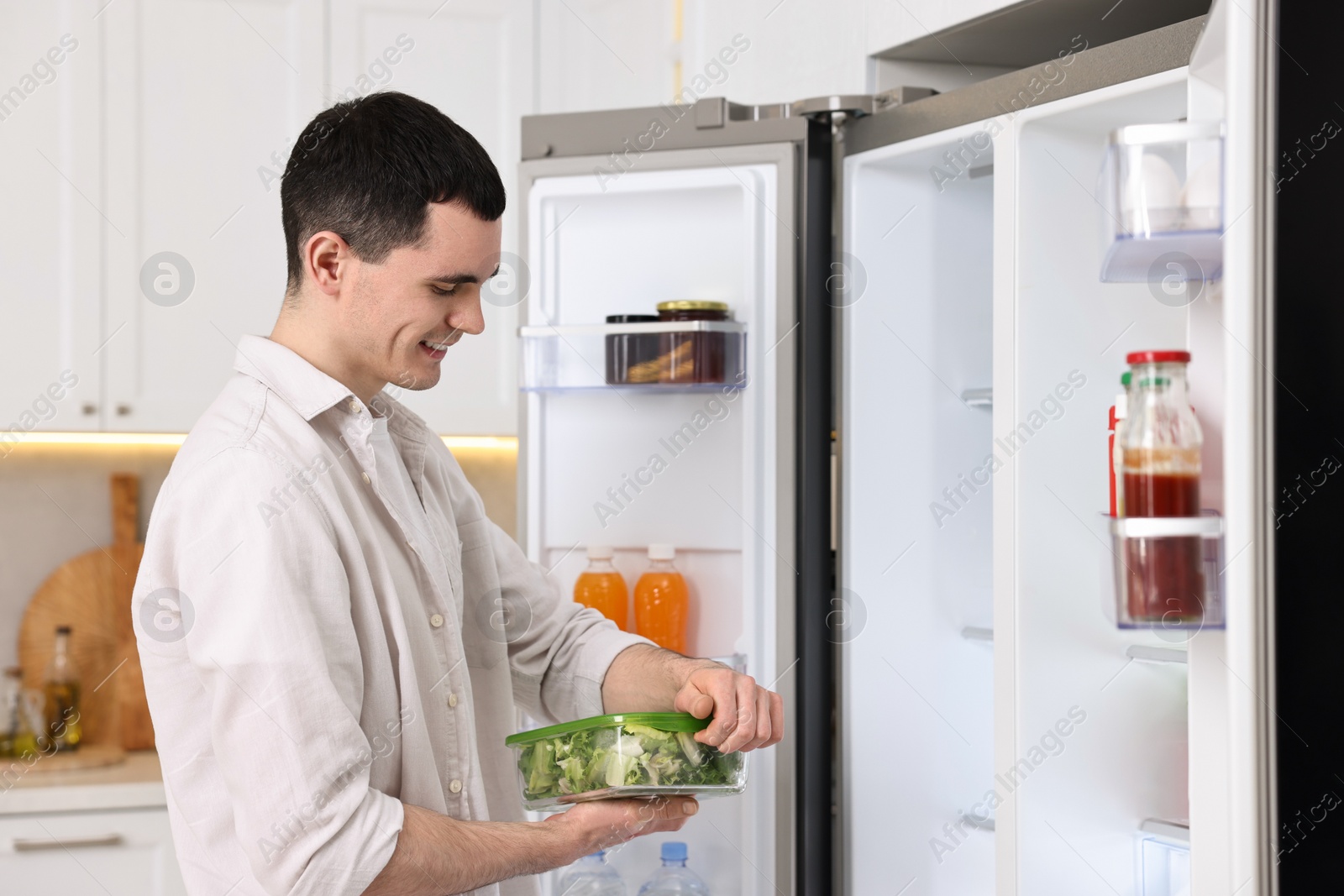 Photo of Happy man holding container with vegetables near refrigerator in kitchen