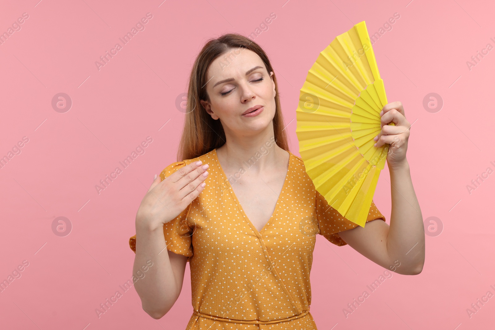 Photo of Beautiful woman waving yellow hand fan to cool herself on pink background