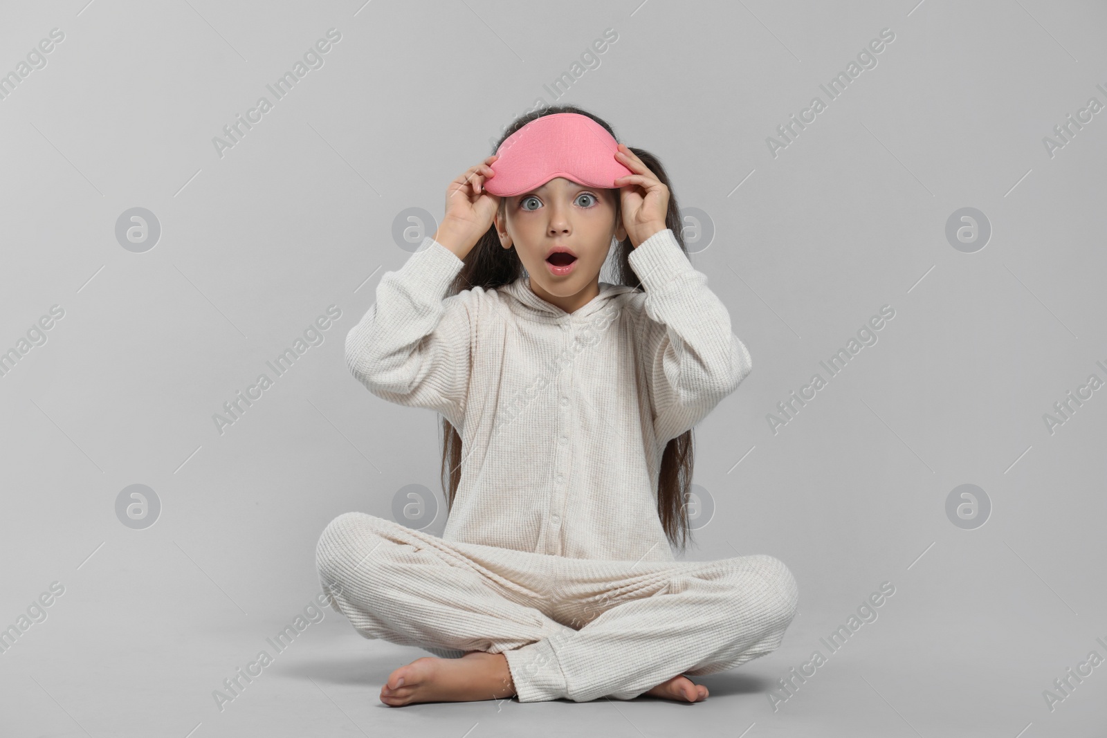 Photo of Surprised girl in white pajamas with pink sleep mask on light grey background