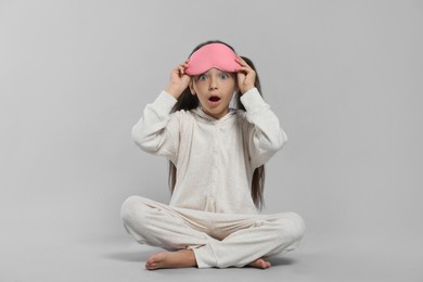 Surprised girl in white pajamas with pink sleep mask on light grey background