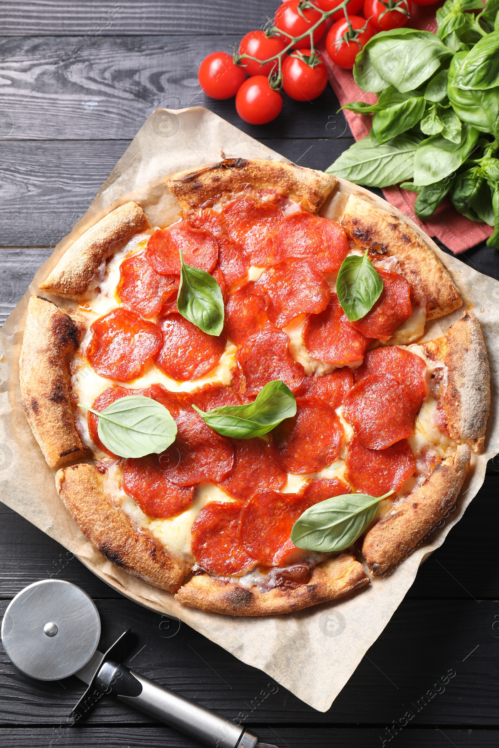 Photo of Tasty pepperoni pizza with basil, tomatoes and cutter on black wooden table, top view