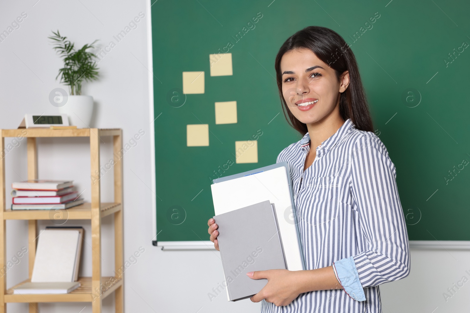 Photo of Happy young teacher with books at blackboard in classroom. Space for text