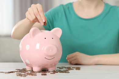 Financial savings. Woman putting coin into piggy bank at white wooden table indoors, closeup