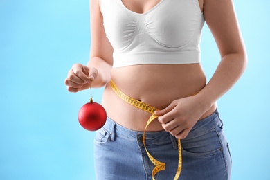 Photo of Woman with Christmas ball and measuring tape on light blue background, closeup. Overweight problem after New Year party