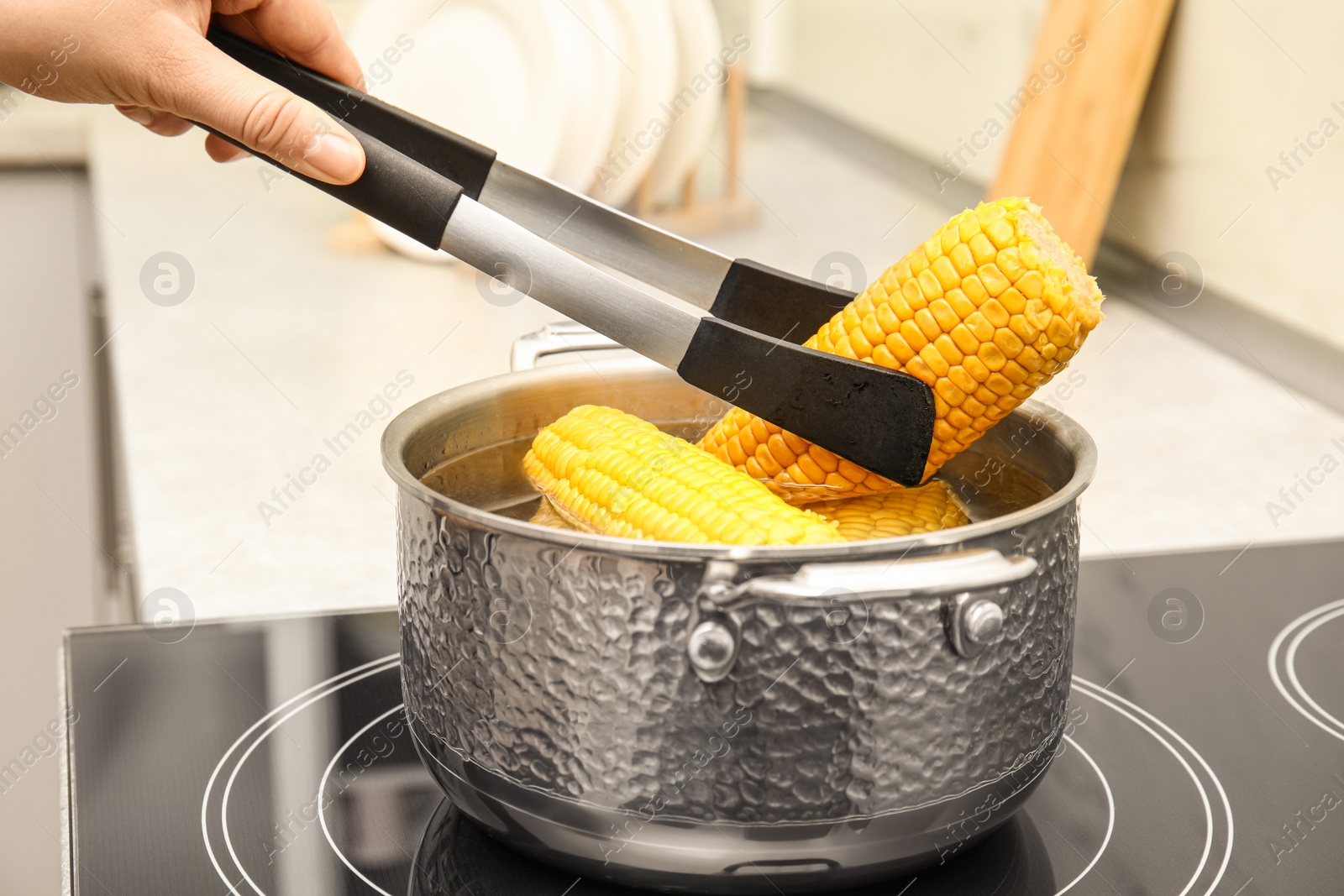 Photo of Woman taking boiled corn from pot with tongs in kitchen, closeup