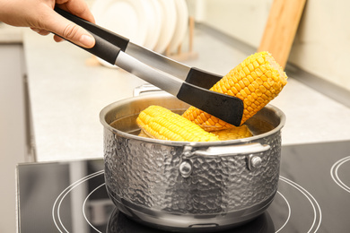 Photo of Woman taking boiled corn from pot with tongs in kitchen, closeup