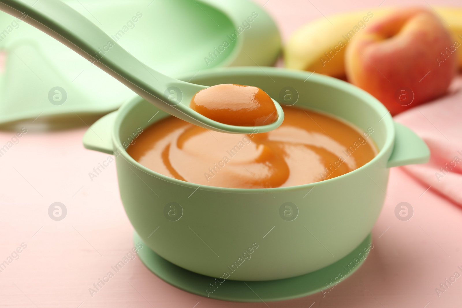 Photo of Spoon with tasty pureed baby food over bowl on pink wooden table, closeup