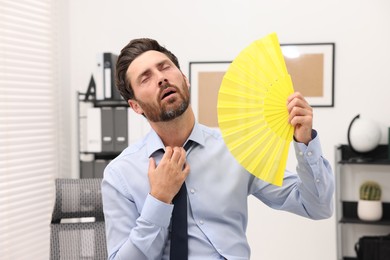 Photo of Bearded businessman waving yellow hand fan to cool himself in office