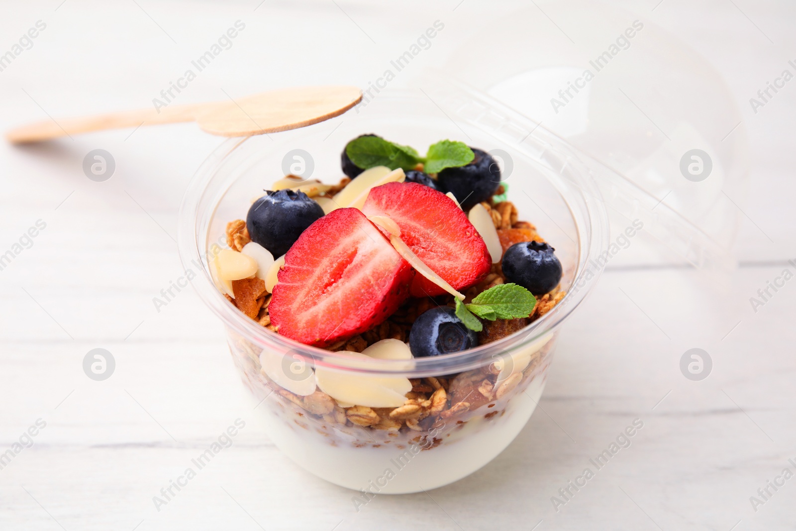 Photo of Tasty granola with berries, almond flakes and yogurt in plastic cup on white wooden table, closeup