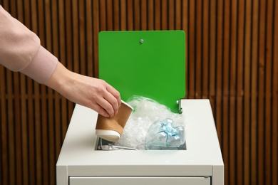 Photo of Woman putting used paper cup into trash bin on wooden background, closeup. Recycling concept