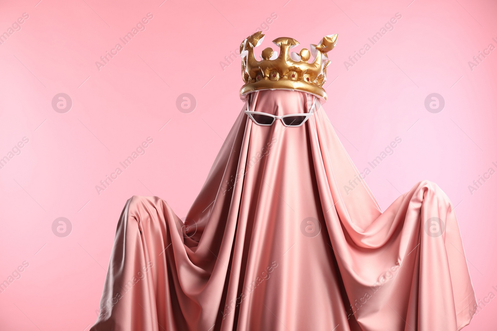 Photo of Glamorous ghost. Woman in sheet with sunglasses and crown on pink background