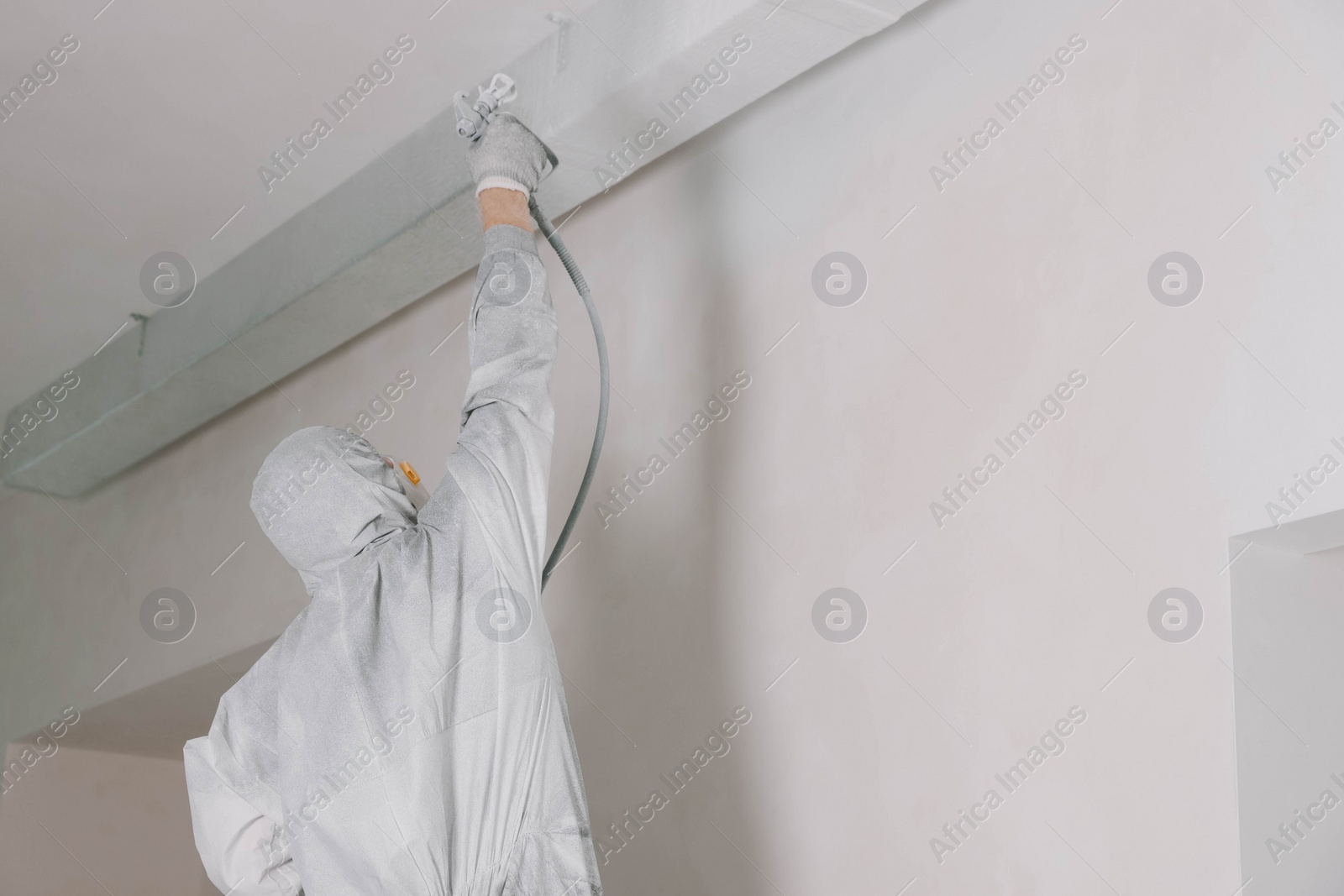 Photo of Decorator in protective overalls painting wall with spray gun indoors