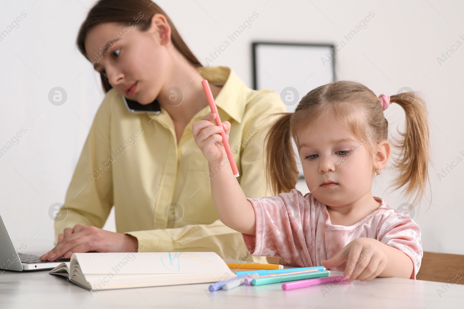 Photo of Woman with laptop working remotely at home. Busy mother talking on phone while her daughter drawing at desk. Selective focus
