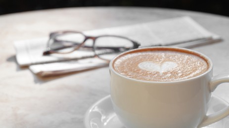 Cup of delicious coffee, glasses and newspaper on beige marble table outdoors, closeup. Space for text