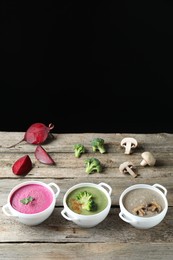 Different tasty cream soups in bowls and ingredients on old wooden table against black background. Space for text