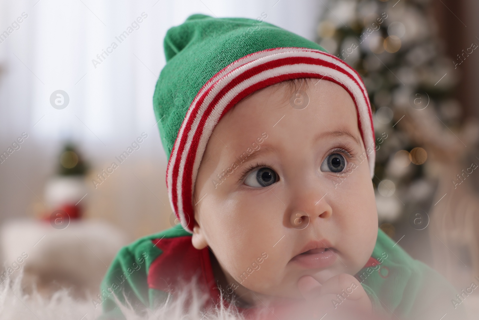 Photo of Baby wearing cute elf costume in room decorated for Christmas