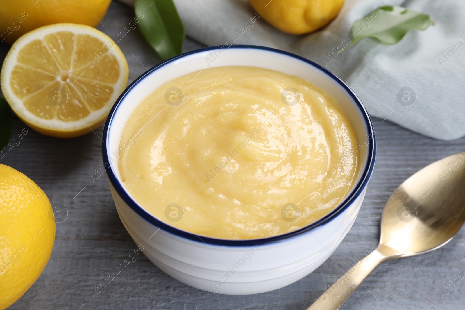 Photo of Delicious lemon curd in bowl, fresh citrus fruits, leaves and spoon on grey wooden table, closeup