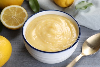 Photo of Delicious lemon curd in bowl, fresh citrus fruits, leaves and spoon on grey wooden table, closeup