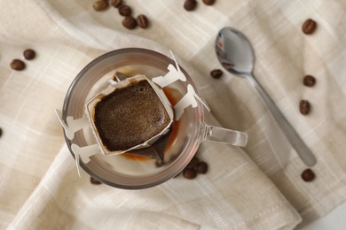 Photo of Glass cup with drip coffee bag and beans on beige fabric, flat lay